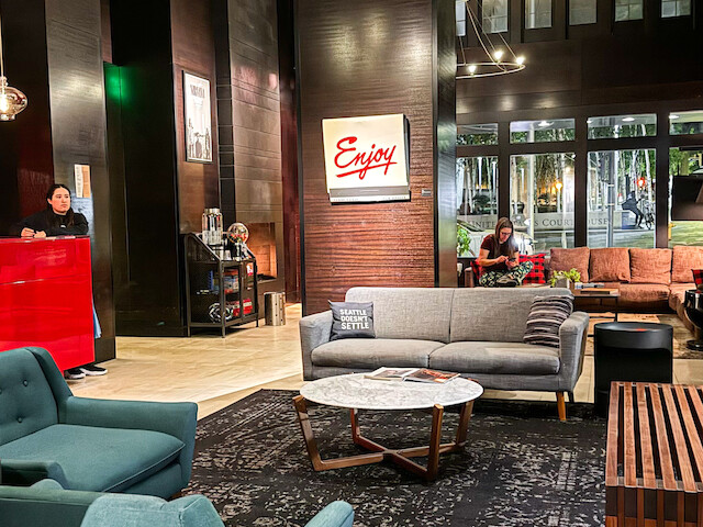 The image shows a modern hotel lobby with seating, a red reception desk, stylish lighting, and decorative elements.