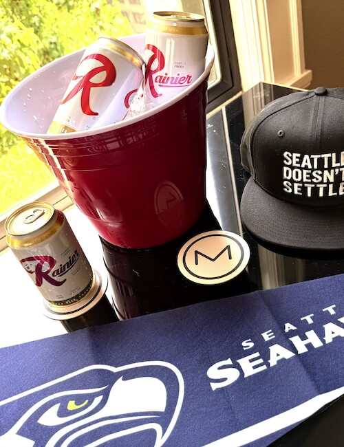 A Seahawks cap, a towel, canned beverages, a bucket with drinks, and a round sticker or coaster with the letter M.