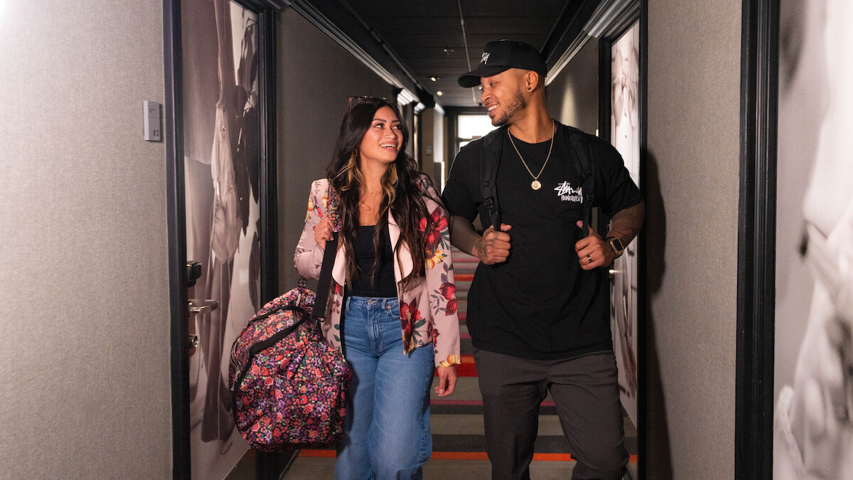 Two young adults walk together down a hallway, smiling at each other and carrying backpacks, suggesting they are friends or travelers.