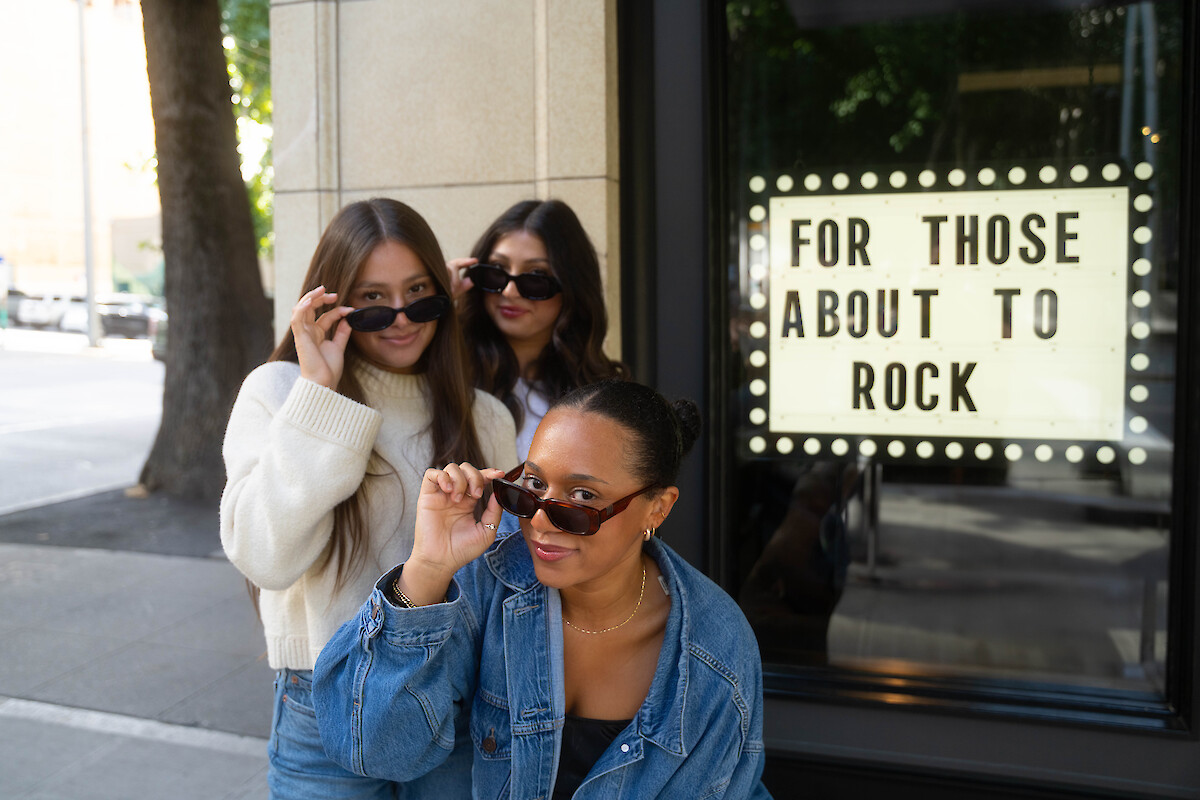 Three women with sunglasses stand outside, playfully posing near a sign that says 